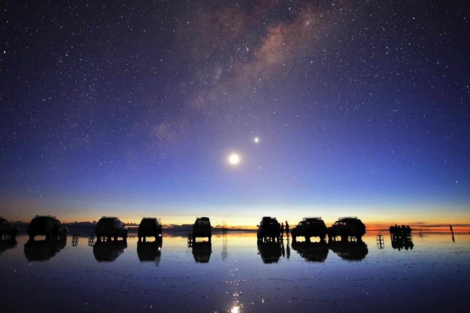 Starry night sky over a reflective water surface with silhouettes of people and 4x4 at Uyuni salt flat, Bolivia.