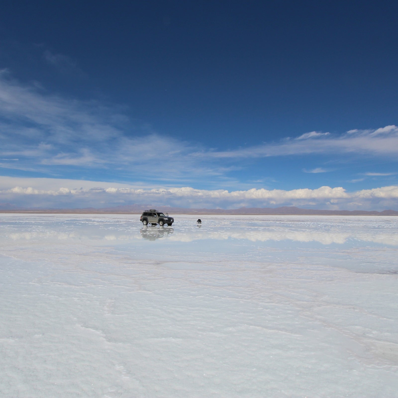 Private 4x4 crossing the wide reflective surface of the Uyuni Salt Flat beneath a deep blue sky.