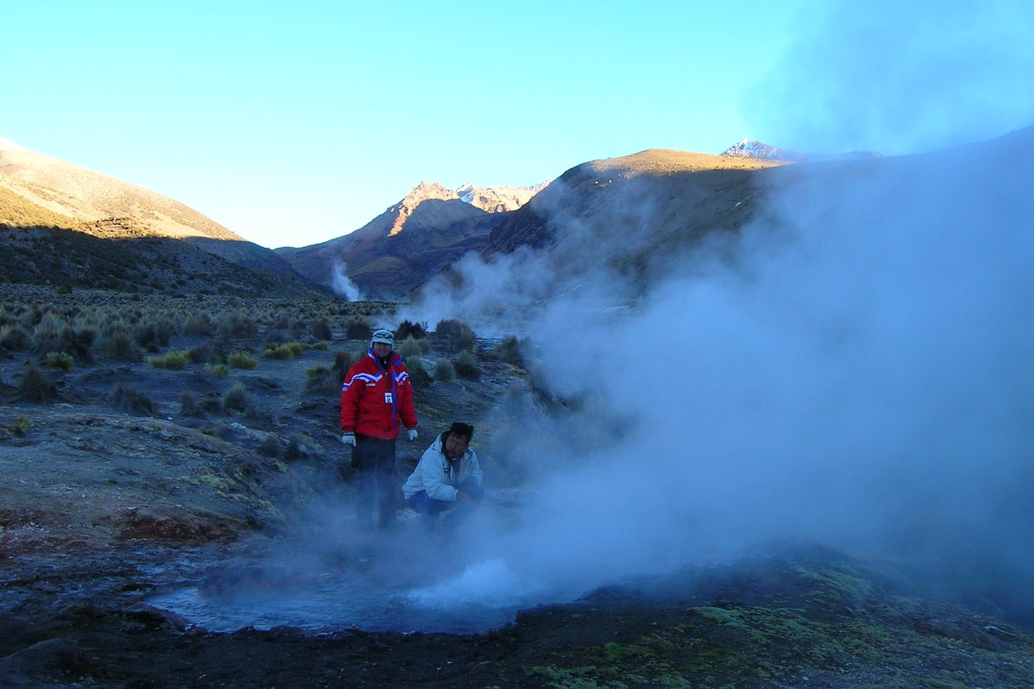 Two people near a geothermal feature with steam rising, surrounded by mountains.