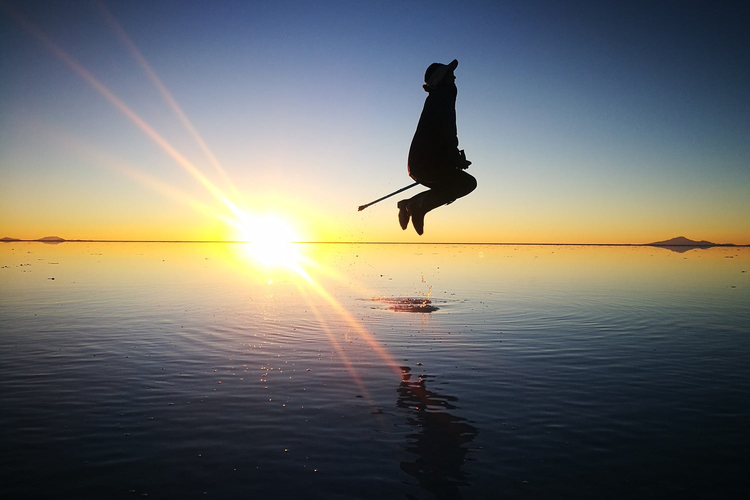 Silhouette of a traveler jumping above the reflective surface of the Uyuni Salt Flat at sunset.
