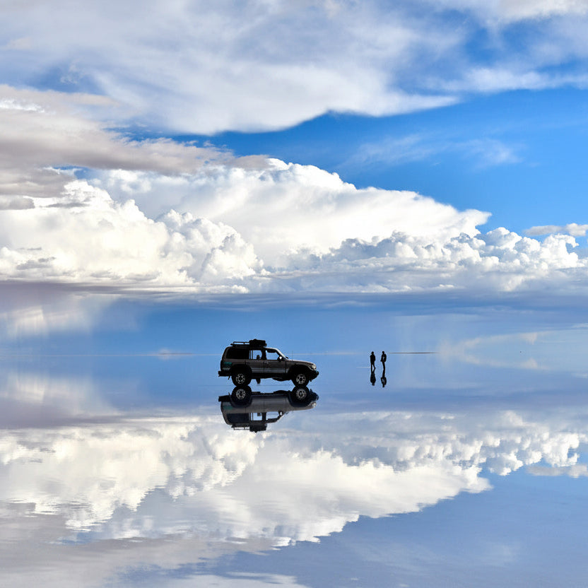 Reflective surface with a car and two people, mirrored against a blue sky with clouds at Uyuni Salt Flats.
