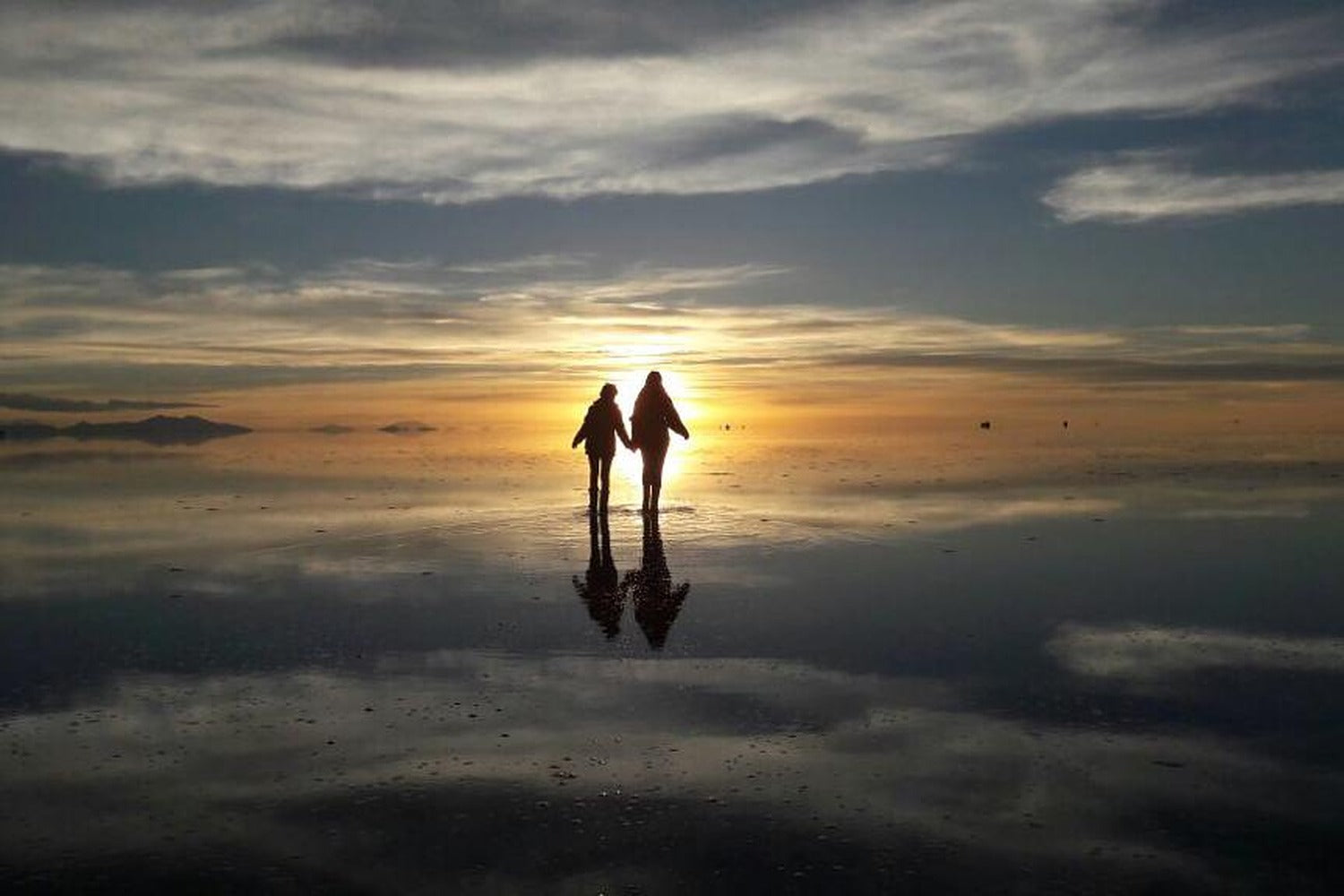 Two travelers holding hands on the reflective surface of the Uyuni Salt Flat at sunset.