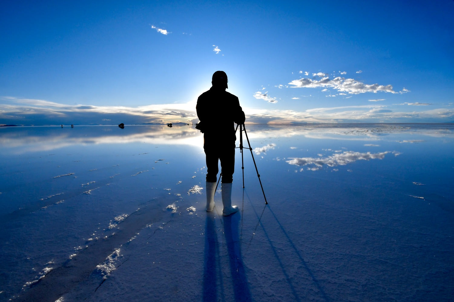 Photographer standing on the reflective Uyuni Salt Flat with tripod, blue sky, and cloud reflections.