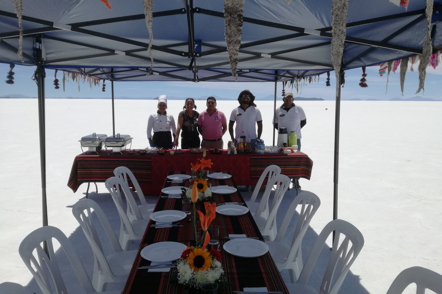 Staff standing around a table with food under a large canopy on a clear day at Uyuni Salt Flats.