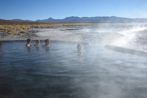 People enjoying a hot spring with mist rising from the water, surrounded by a desert landscape.