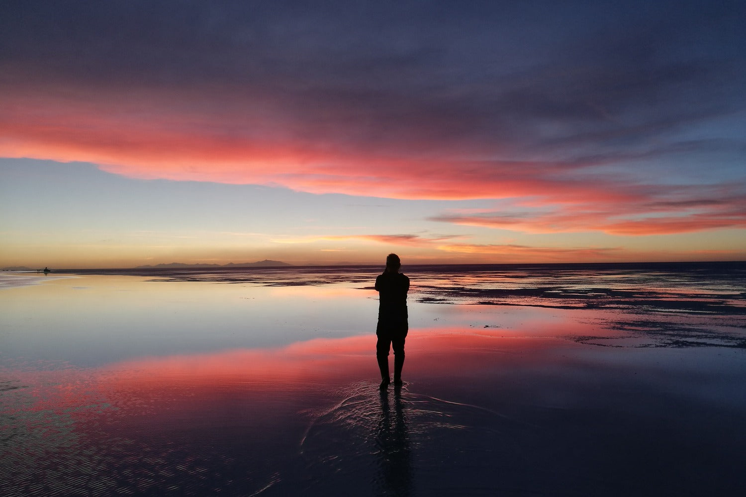 Silhouette contemplating a spectacular sunset over the Uyuni Salt Flats, where the vivid colors of the sky reflect on the water creating a scene of serenity and unforgettable beauty.