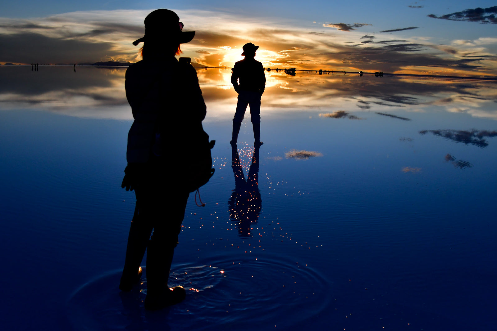 Two travelers standing on the reflective Uyuni Salt Flat at sunset.