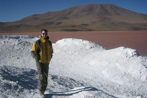 Person posing in front of a stunning high-altitude lagoon with reddish waters, surrounded by volcanic landscapes and desert mountains in the South Lipez