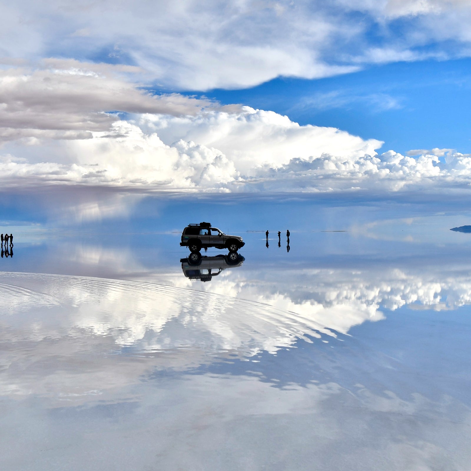 4x4 vehicle crossing the spectacular mirror effect of the Uyuni Salt Flats, where the sky and clouds reflect perfectly.