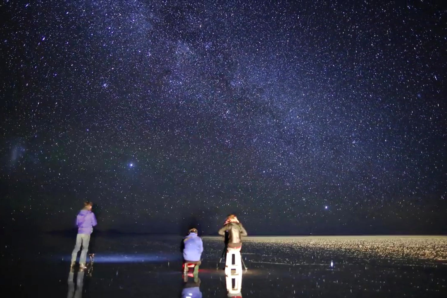 Three people observing the night sky filled with stars over Uyuni salt flat reflective surface.