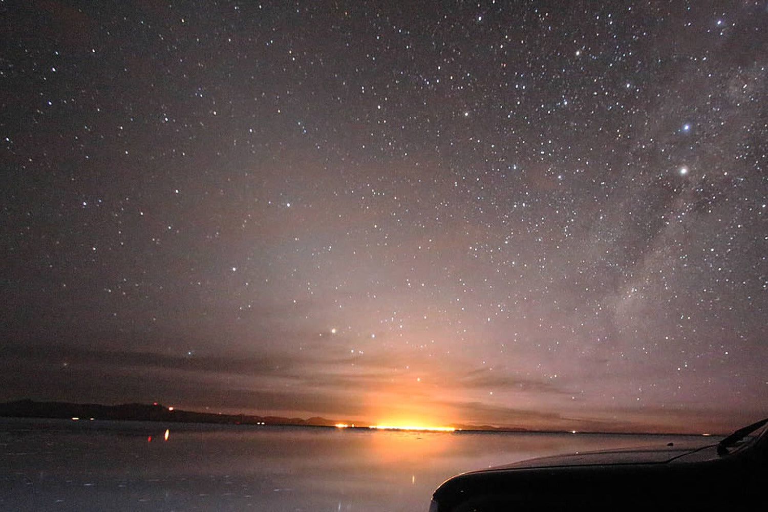 Starry night sky over Uyuni salt flat water with a Uyuni town  light in the distance.